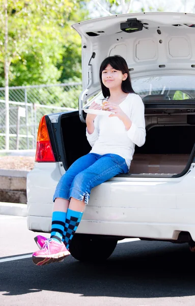 Preteen girl sitting on back car bumper eating lunch — Stock Photo, Image