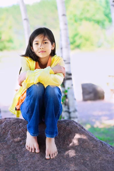 Young biracial girl sitting on rock under trees — Stock Photo, Image