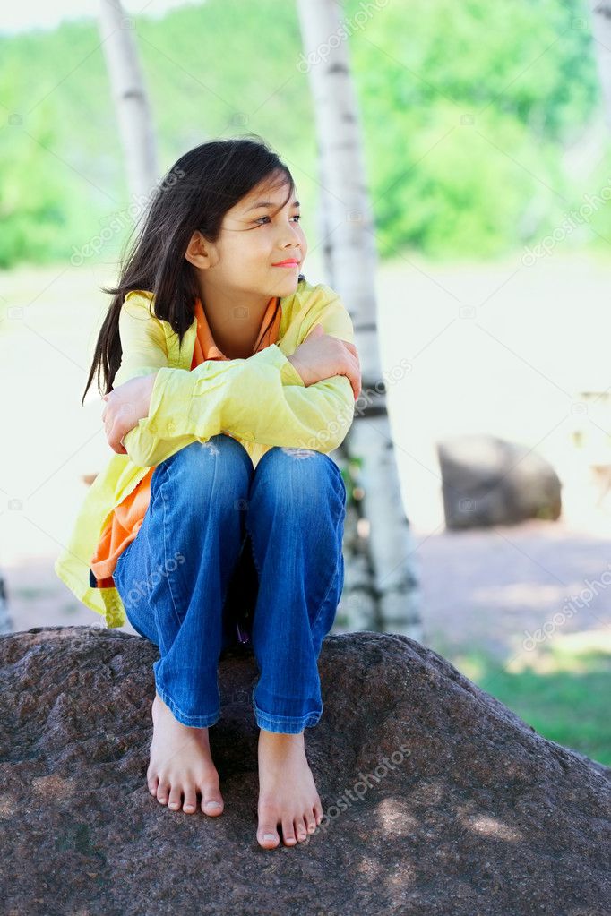 Young biracial girl sitting on rock under trees