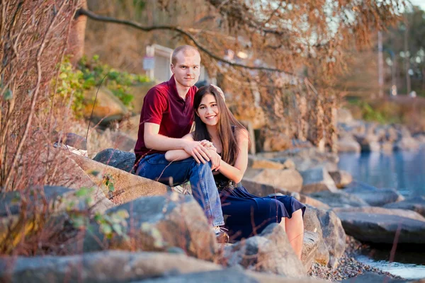 Young interracial couple sitting together on rocky shoreline by — Stock Photo, Image