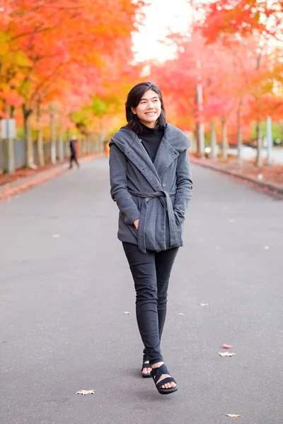 Smiling teen girl in gray jacket walking along road enjoying colorful autumn leaves
