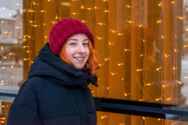 Pencerenin yanındaki genç ve güzel bayan kışın City Caddesi 'nde Noel çelengiyle süslenmiş. Genç kız akşamları cam vitrine bakıyor. Şehir tatili, kış tatili, şehir konseptinde tatiller..