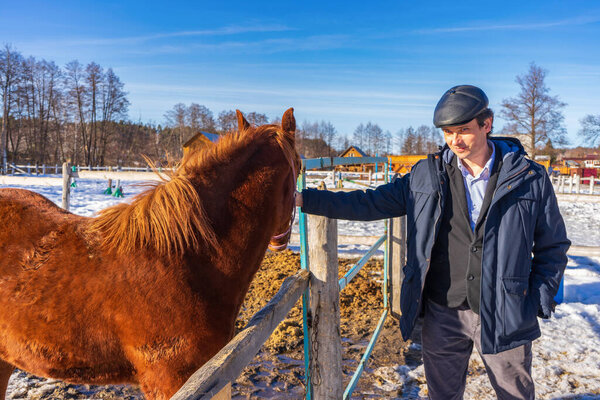 Farmer man with horse at ranch in winter sunny day. Owner and his animal. Winter weekend at farm, trip to countryside. Healthy lifestyle, active leisure, authentic moments.