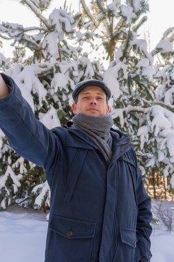 Güneşli bir günde karla kaplı çam ağaçlarıyla kış manzarasına selfie çeken orta yaşlı yakışıklı bir adamın portresi. İnsan ve doğa, kırsal alanda kış haftasonu konsepti.