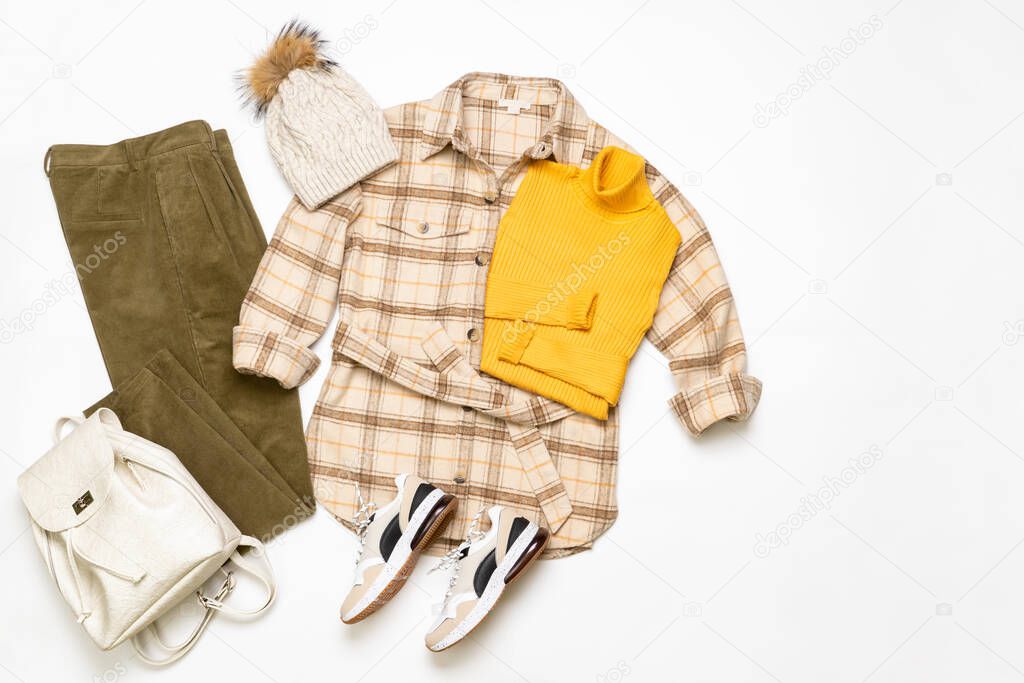 Female winter or autumn stylish clothing set. Plaid checkered shirt, yellow sweater, green corduroy trousers, hat with pompom, sneakers and backpack. Trendy fashionable casual clothes. Fashion