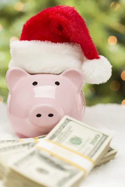 Roze piggy bank dragen KERSTMUTS in de buurt van stapels van geld op snowfl — Stockfoto