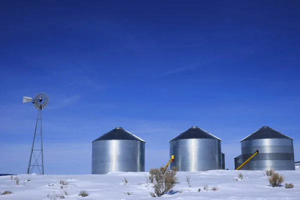 Windmill and steel grain silos in winter snow with blue sky on farm for agricultural farming