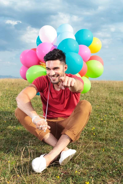 Seated casual man with balloons points at you — Stock Photo, Image
