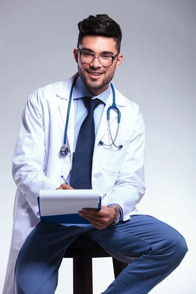 Seated young doctor writing on clipboard — Stock Photo, Image