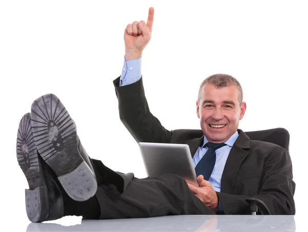 Hombre de negocios con los pies en el escritorio sostiene su tableta y señala hacia arriba — Foto de Stock