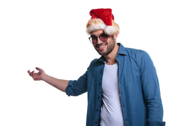 Giovane uomo casual in cappello di Babbo Natale presenta qualcosa — Foto Stock