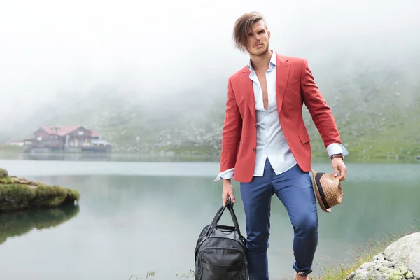 Hombre de moda sosteniendo su sombrero y bolsa de viaje cerca del lago — Foto de Stock