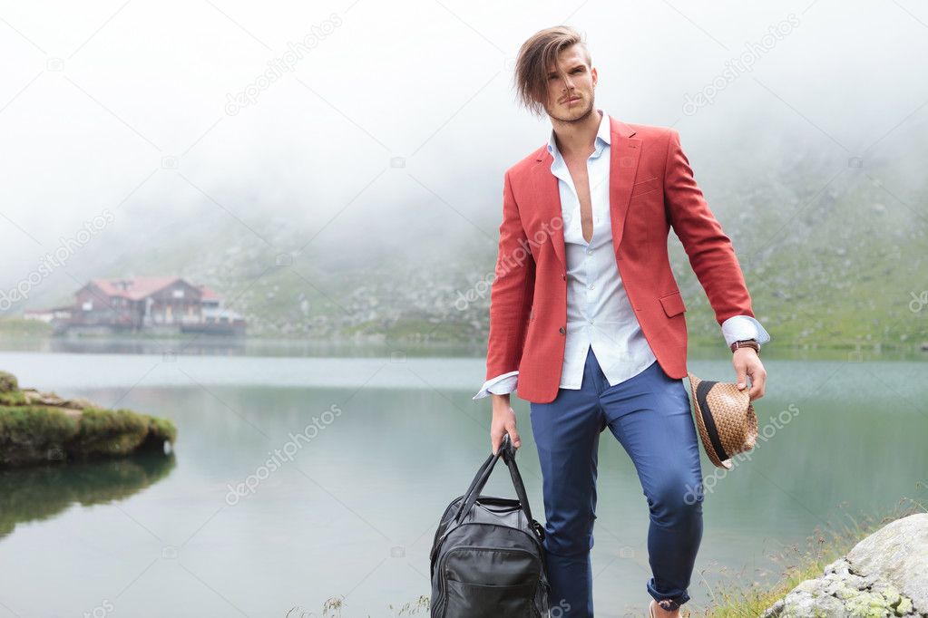 fashion man holding his hat and travel bag near lake
