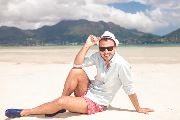 Мужчина сидит на пляже и держит шляпу. — стоковое фото
