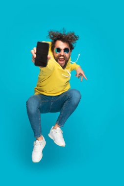 sarı kapüşonlu dinamik genç adam siyah boş ekranı gösteriyor havaya zıplıyor ve stüdyoda mavi arka planda gülüyor.