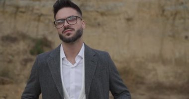 Gözlüklü, kendine güveni tam sakallı bir adam yukarı bakıyor. Gri ceketini ilikleyip dışarı bakıyor ve poz veriyor.