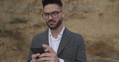 Tıraşsız, gri ceketli, gözlüklü mutlu bir adam. Doğanın dışında vakit geçiriyor. Telefonu tutuyor, mesaj atıyor, gülümsüyor ve göz kırpıyor.