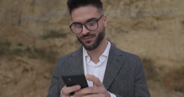 灰色のジャケットに身を包んだ剃らないクールな男は 眼鏡をかけ 電話をかけ テキストメッセージをかけ 微笑み うなずき そして自然の中で外にウィンク — ストック動画