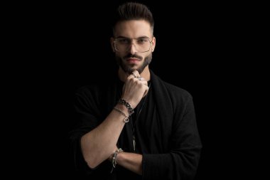 Sakallı, gözlüklü, çenesine dokunup düşünen, kollarını kavuşturup siyah arka planda poz veren yakışıklı bir genç.