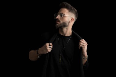 Gözlüklü, kendine güvenen sakallı genç adam siyah arka planda poz verirken ceketini tutuyor ve düzenliyor.