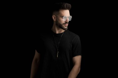 Havalı sakallı adam gözlük takıyor ve siyah arka planda poz veriyor.