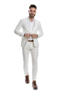 Seksi manken takım elbisesini kapatıyor, yürüyor ve beyaz stüdyo arka planında kameraya bakıyor.