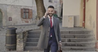 Uzun paltolu sakallı genç adam bavulunu tutuyor, telefonla konuşuyor ve ortaçağ şehrinin önünde yürüyor.