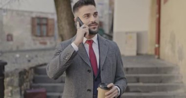 Takım elbiseli işadamı telefonda konuşuyor, elinde kahve tutuyor, kaldırıyor, gülümsüyor ve yürüyor.