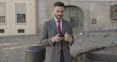 Takım elbiseli, uzun paltolu çekici bir genç. Elinde telefon, kaydırma, kahkaha ve kahkaha ile ortaçağ arka planında kahve içiyor.