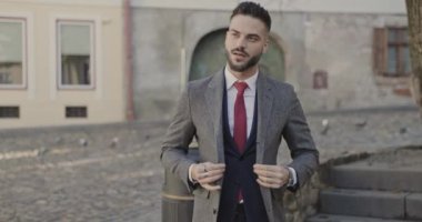 Takım elbiseli zarif, tıraşsız bir iş adamı uzun bir palto hazırlayıp düğmelerini ilikliyor ve ortaçağ kasabasının dışında gülümsüyor.