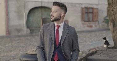 Uzun paltolu çekici sakallı adam, kente bakıyor, kravatını düzeltiyor, gülümsüyor ve ortaçağ arka planında poz veriyor.