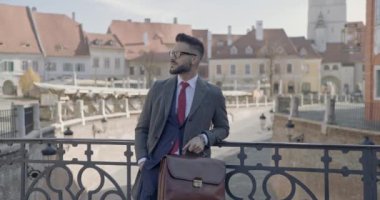 Seksi iş adamı bir eli cebinde, diğer elinde evrak çantasıyla çitlere yaslanıyor, gözlük takıyor ve tavır takmaya çalışıyor.