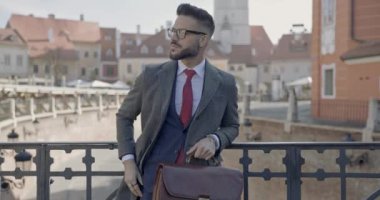 Çekici iş adamı çantasını kapatıyor, bir elini cebine sokuyor, gözlüklerini tamir ediyor ve gülümseyerek parmaklarını şaklatıyor.