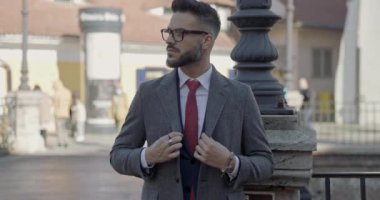 Takım elbiseli seksi bir genç adam, gözlük takıyor, uzun bir palto giyiyor ve düğmelerini ilikliyor, gülümsüyor ve ortaçağ şehrinin dışında bir tarafa bakıyor.