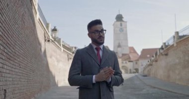 Uzun paltolu kendine güvenen genç adam avuçlarını ovalıyor, parmaklarına dokunuyor, gülümsüyor ve ortaçağ arka planında ceketini ayarlıyor.