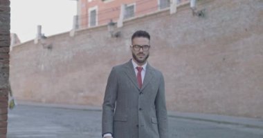 Tıraşsız, çekici, takım elbiseli, uzun paltolu, bavulu tutan, yan tarafa bakıp uzaklaşan bir adam.