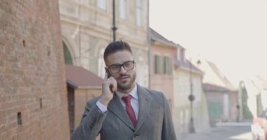 Uzun paltolu ve gözlüklü zarif bir adam telefonda konuşuyor, gülümsüyor ve ortaçağ şehir arka planında yürüyor.