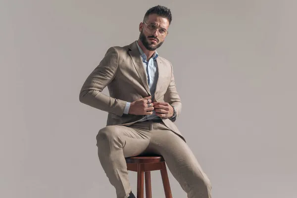 セクシーなファッションビジネスマンが椅子に態度でポーズをとってグレーの背景にジャケットを固定する — ストック写真