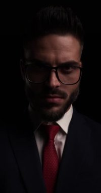 Dramatik traşsız iş adamı, gözlükleriyle aşağıya bakıyor, gülümsüyor, sessiz jest yapıyor, yana bakıyor ve karanlıkta kayboluyor.
