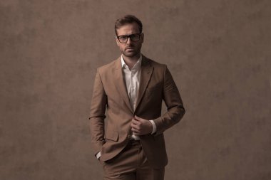 cool sexy businessman holding one hand on his button and the other in his pocket against brown background
