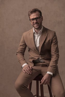 happy businessman smiling at the camera, resting on a chair and posing against brown background