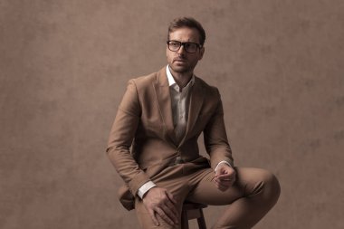 cool sexy businessman resting on a chair, looking away and wearing eyeglasses in a fashion pose
