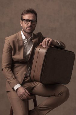 sexy fashion businessman with attitude is posing on a chair holding his briefcase and wearing eyeglasses
