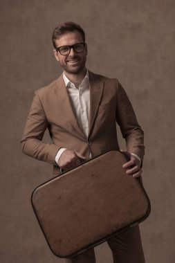 happy businessman holding his briefcase, smiling and wearing eyeglasses on brown background