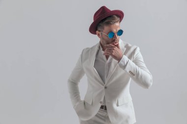 cool fashion model sensually touching his chin, wearing a burgundy hat and sunglasses, holding one hand in pocket