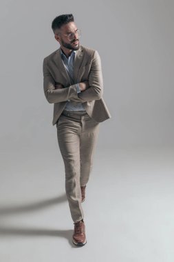 cool young model in suit with glasses looking to side and crossing arms while walking on grey background in studio