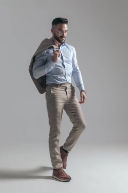 attractive young man with jacket over shoulder looking to side and posing while walking on grey background in studio