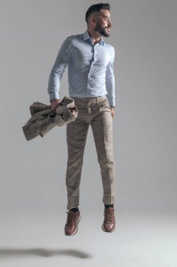 sexy young guy with glasses looking to side, holding jacket in hand and jumping on grey background in studio