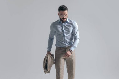 portrait of attractive young man with glasses looking down and holding jacket while walking on grey background in studio