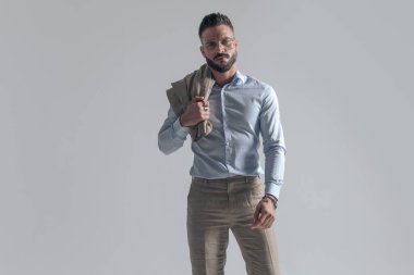 sexy unshaved guy holding jacket over shoulder, wearing glasses and posing in a confident manner on grey background in studio